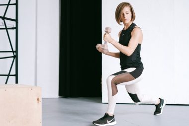 A young woman performing lunges with dumbbells in a modern indoor gym setting.